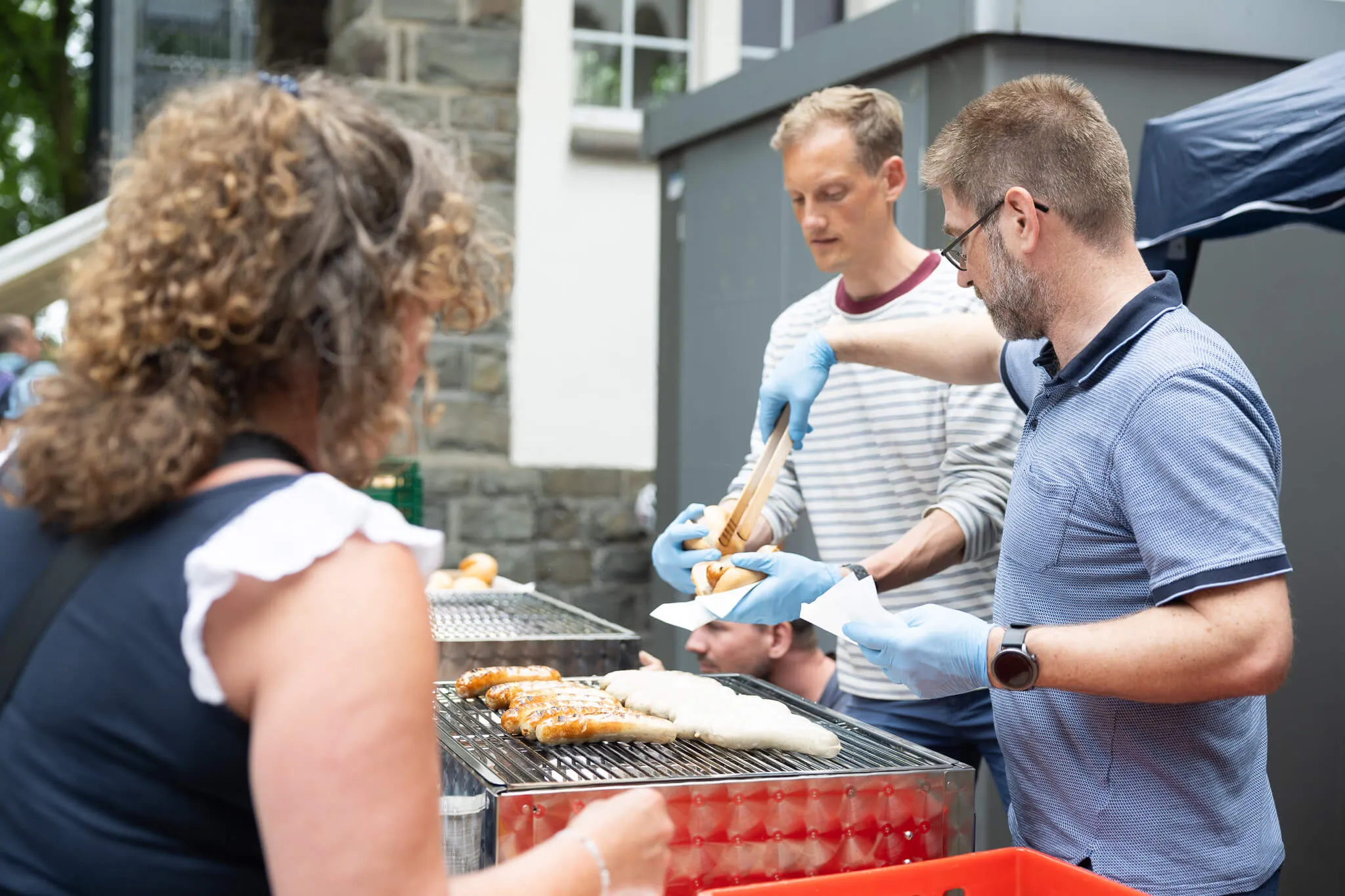 Zwei Männer mit blauen Handschuhen grillen Würstchen auf einem mobilen Grill, während eine Frau mit gelocktem Haar eine Wurst entgegennimmt. Im Hintergrund steht ein weiteres Kind und ein Mann beobachtet das Geschehen.