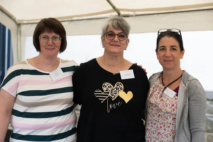 Drei lächelnde Frauen stehen nebeneinander vor einem weißen Zelt. Jede trägt ein Namensschild.
