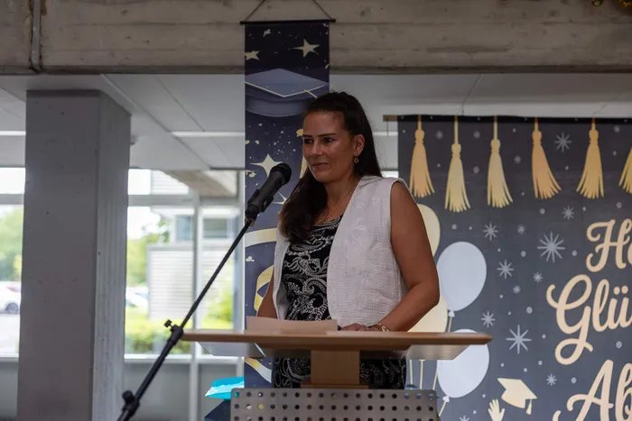 Eine Frau spricht an einem Rednerpult vor einem Publikum. Im Hintergrund eine Dekoration mit Abschlussmützen und der Schriftzug „Herzlichen Glückwunsch“.