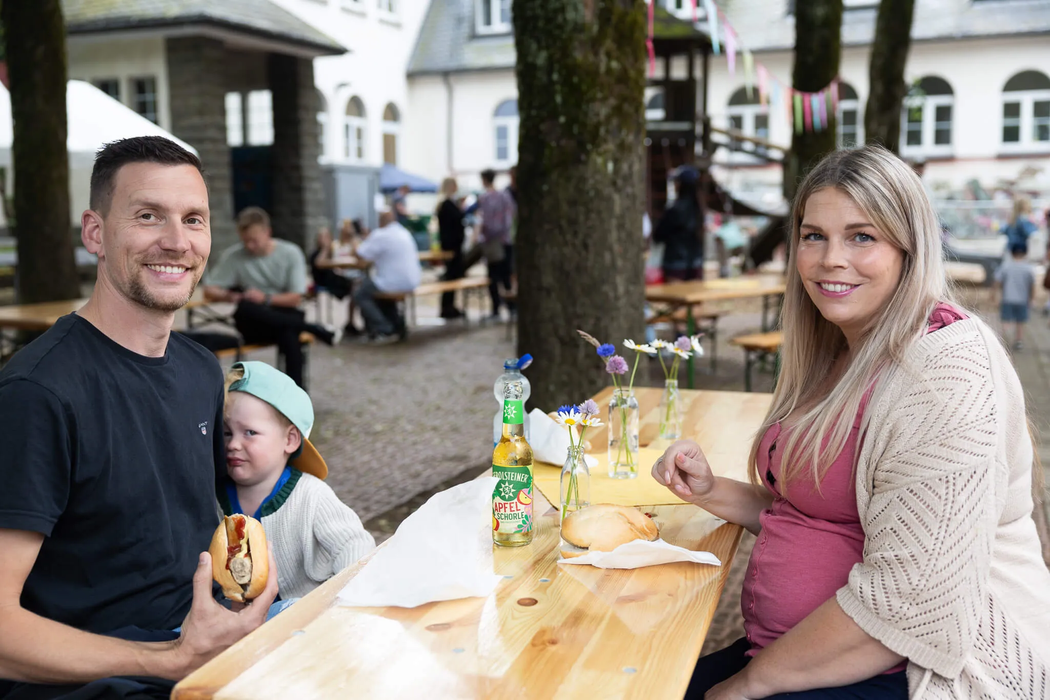 Familie sitzt an einem Holztisch vor der Lindenhofschule und isst. Im Hintergrund sind weitere Besucher des Schulfestes zu sehen.