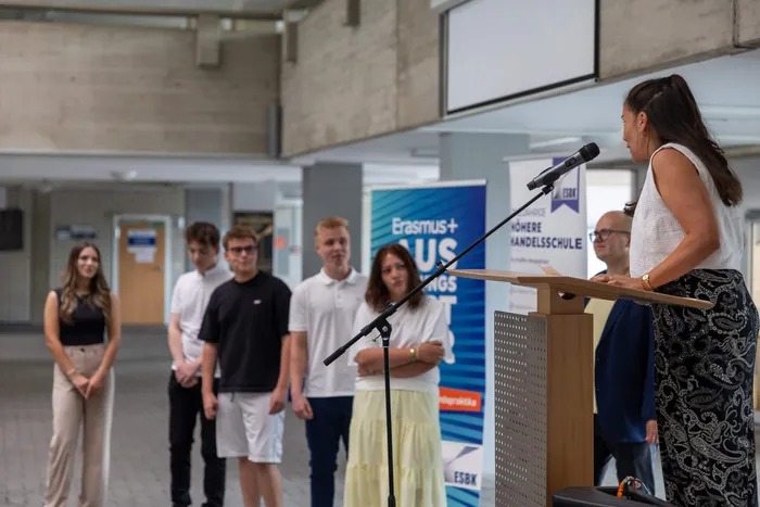 Eine Frau spricht an einem Pult vor einer Gruppe von jungen Menschen, darunter zwei Mädchen und drei Jungen. Im Hintergrund ein Banner mit der Aufschrift „Erasmus+“ und dem Logo der ESBK, der Höheren Handelsschule.