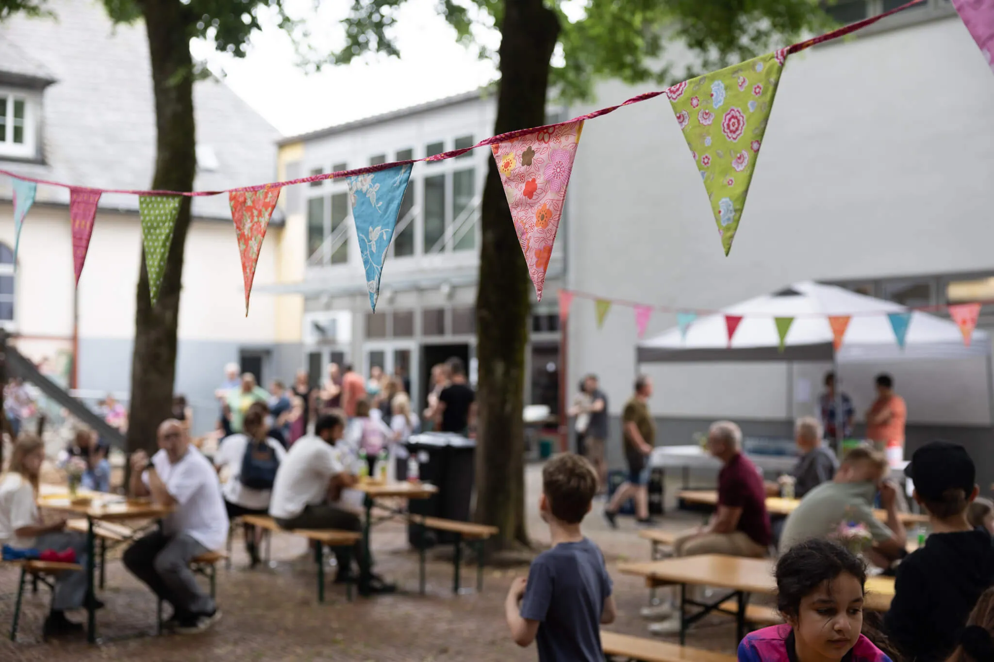 Menschen sitzen an Biertischgarnituren vor einem Gebäude mit Arkaden und feiern auf dem Schulhof der Lindenhofschule. Bunte Wimpelketten hängen zwischen den Bäumen.