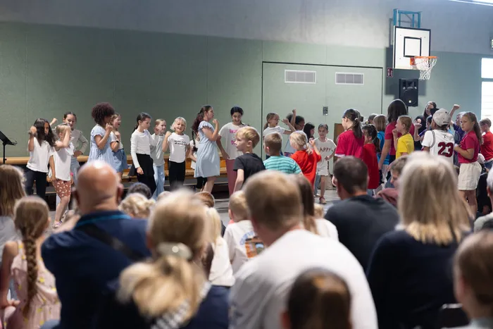Eine Gruppe von Kindern und Jugendlichen steht auf einer Bühne in einer Turnhalle, umgeben von Zuschauern. Im Hintergrund ein Basketballkorb und grüne Wände.