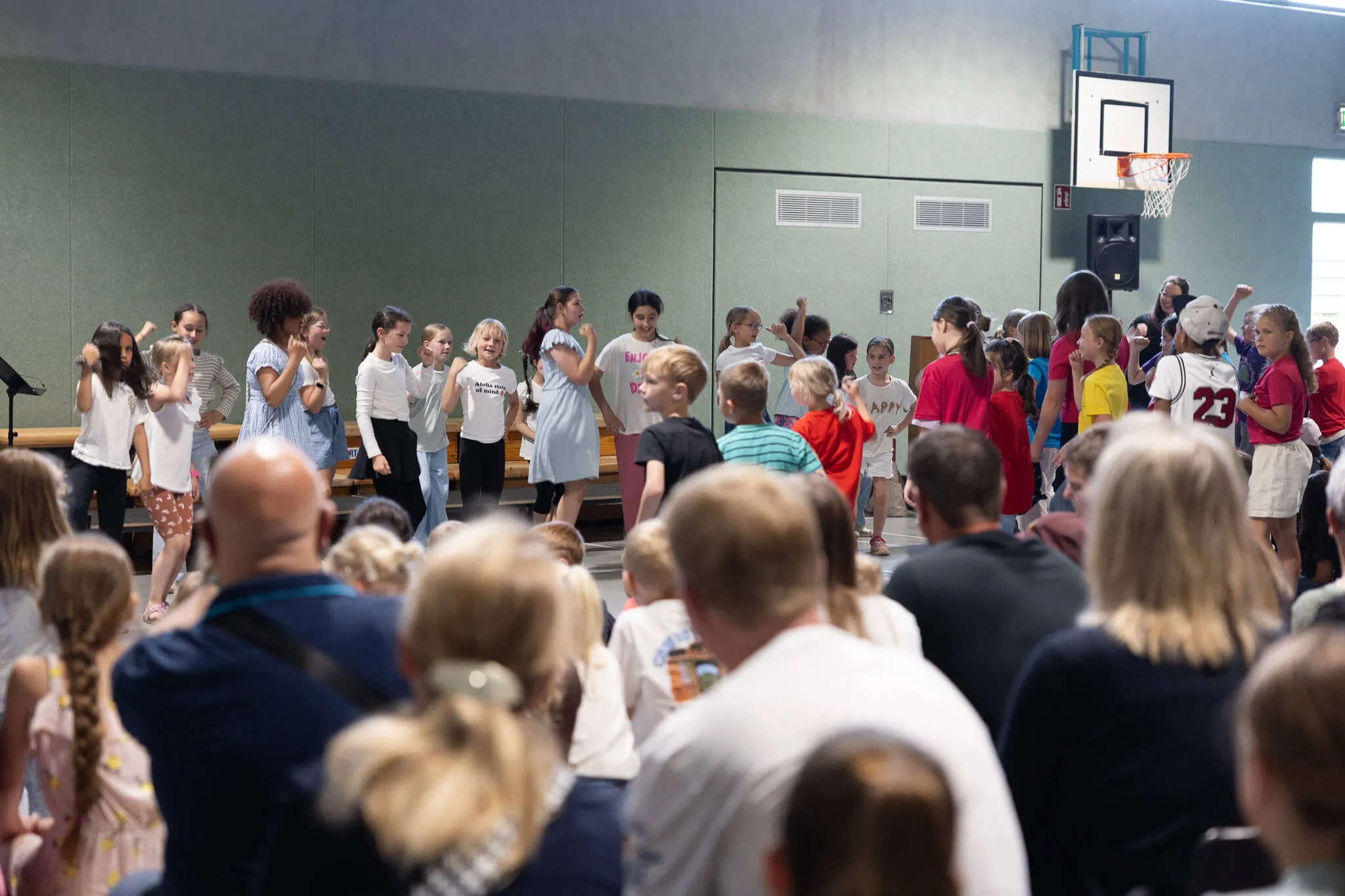 Eine Gruppe von Kindern und Jugendlichen steht auf einer Bühne in einer Turnhalle, umgeben von Zuschauern. Im Hintergrund ein Basketballkorb und grüne Wände.