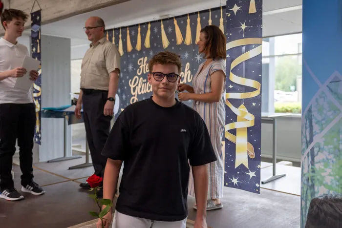 Ein junger Mann mit roter Brille und schwarzem T-Shirt hält eine rote Rose im Blickfeld, im Hintergrund stehen Menschen vor einer mit Glückwünschen geschmückten Wand.