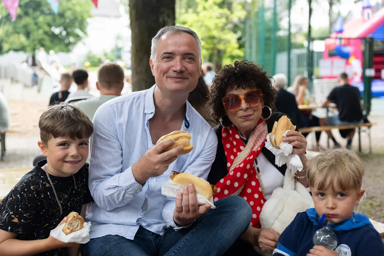 Familie mit zwei Kindern hält jeweils ein belegtes Brötchen in der Hand und lächelt in die Kamera. Im Hintergrund sind weitere Menschen und ein buntes Schulfest zu sehen.