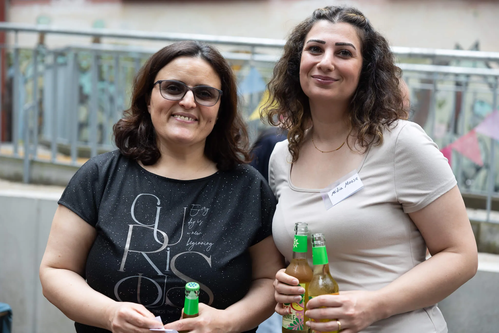 Zwei lächelnde Frauen stehen eng nebeneinander und halten Bierflaschen. Im Hintergrund sind weitere Menschen und ein Gebäude zu sehen.