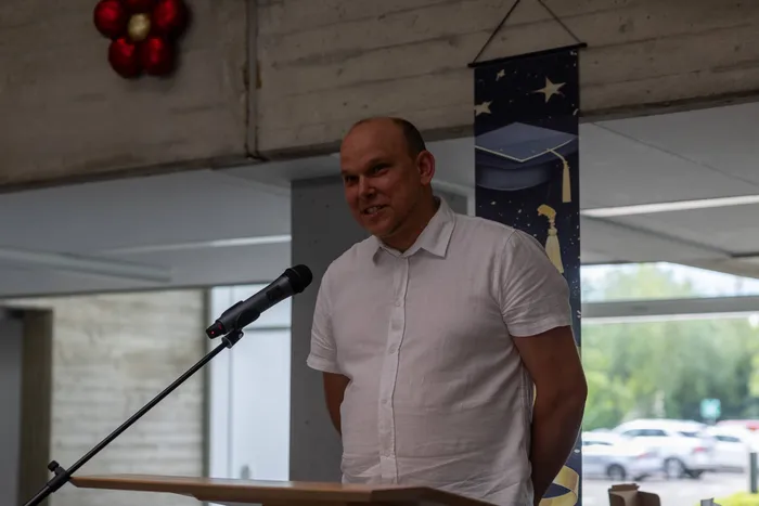Ein Mann mit Glatze und weißem Hemd spricht in ein Mikrofon an einem Rednerpult. Im Hintergrund ein Banner mit einem Diplomhut und Sternen sowie ein Blick durch ein Fenster auf parkende Autos.