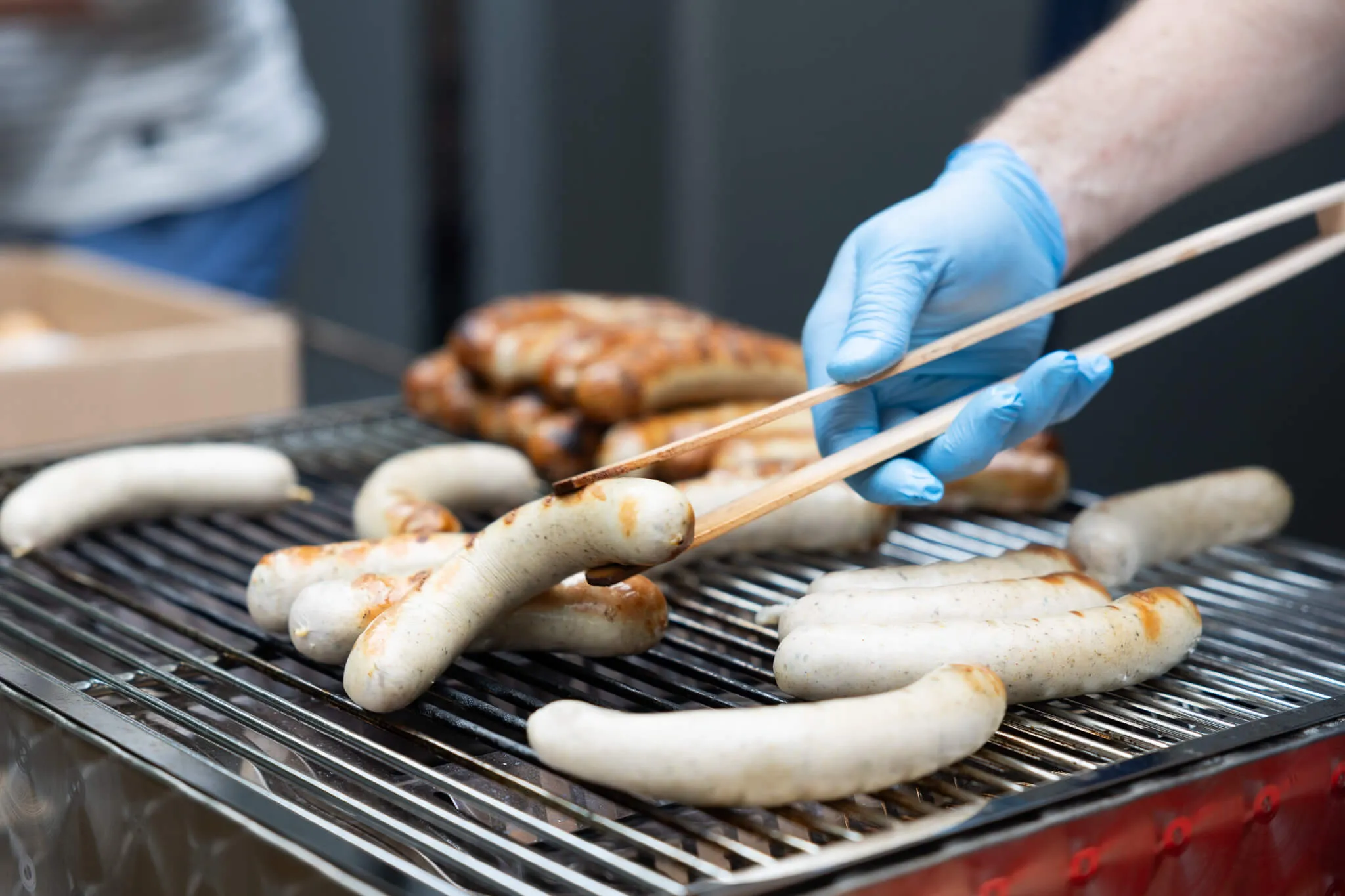 Eine Person mit blauen Handschuhen wendet Bratwürste auf einem Grill mit einer Zange.