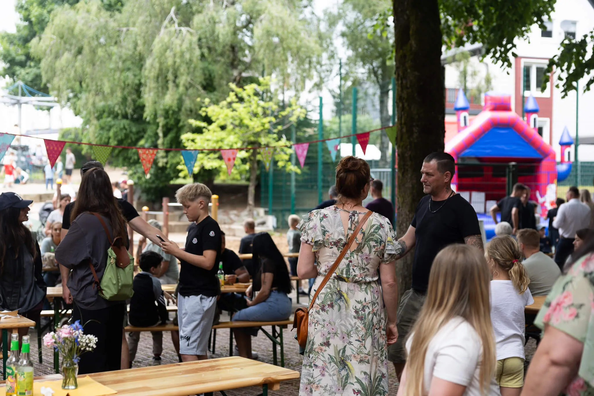Menschen stehen und sitzen an Biertischbänken auf dem Gelände der Lindenhofschule während eines Schulfestes. Im Hintergrund ist ein Hüpfschloss und Girlanden zu sehen.
