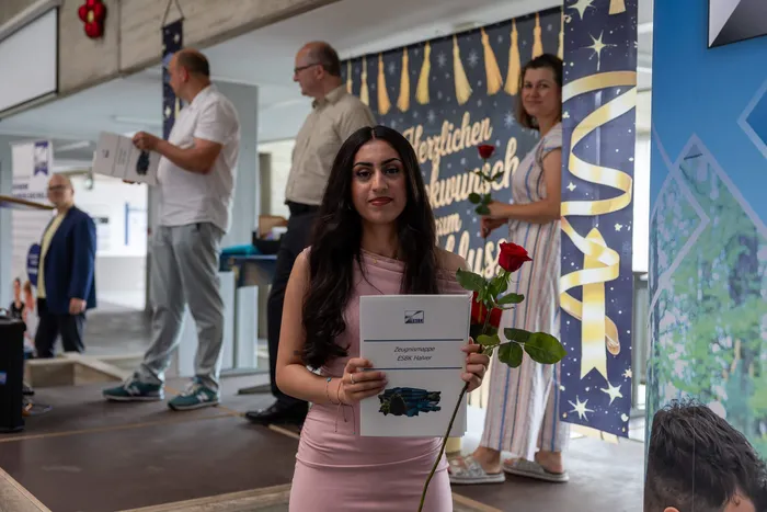 Eine junge Frau mit dunklen Haaren hält eine Urkunde und eine rote Rose. Im Hintergrund sind weitere Personen und ein Banner mit der Aufschrift 