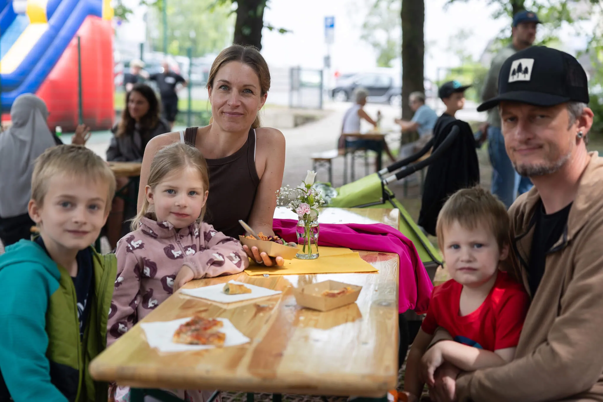 Familie sitzt an einem Tisch mit Pizza und Snacks während eines Schulfestes im Freien. Im Hintergrund sind weitere Menschen und eine Hüpfburg zu sehen.