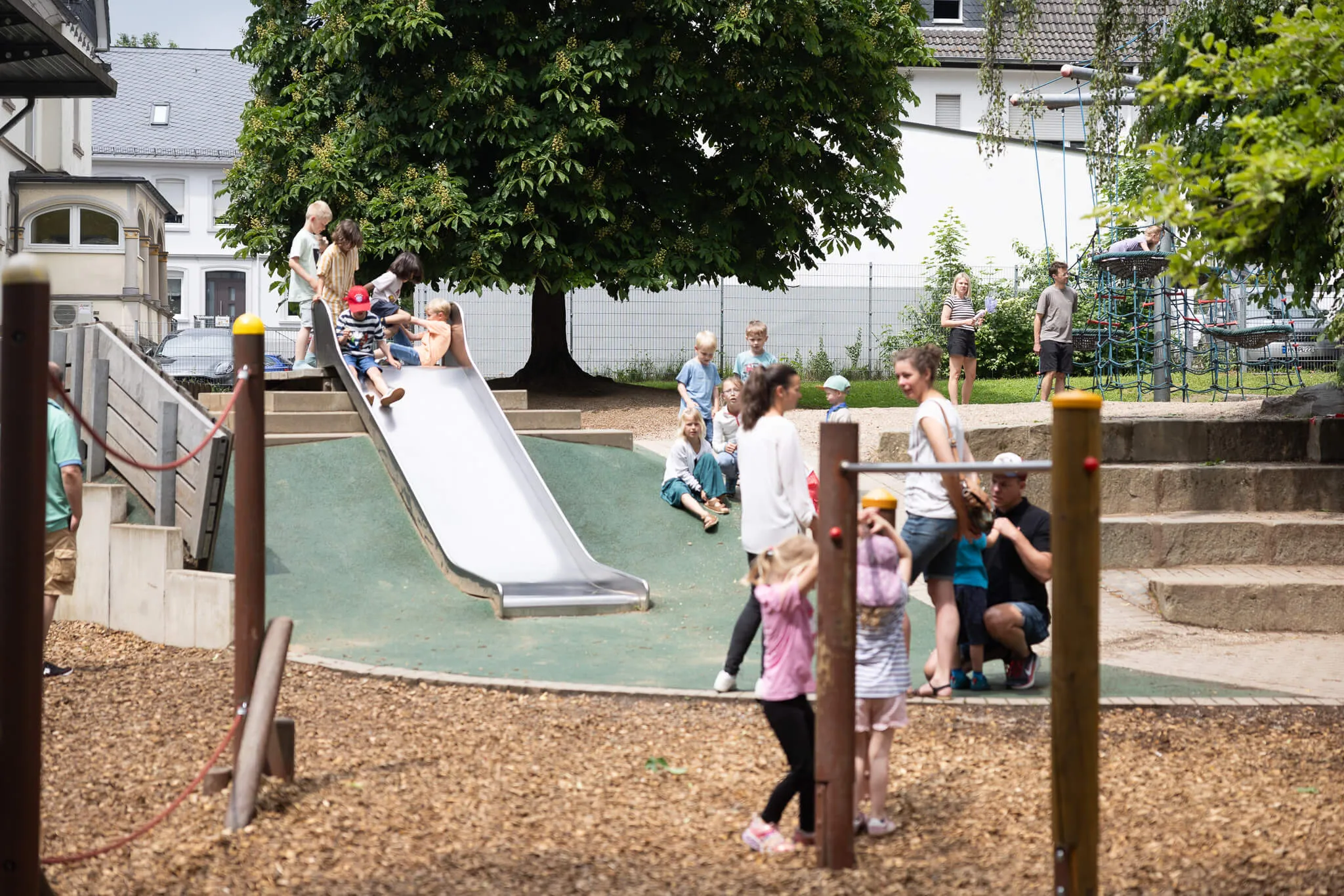 Kinder rutschen auf einer Metallrutsche auf dem Schulhof, während andere spielen und sich unterhalten. Im Hintergrund ist ein Musikschulgebäude zu sehen.