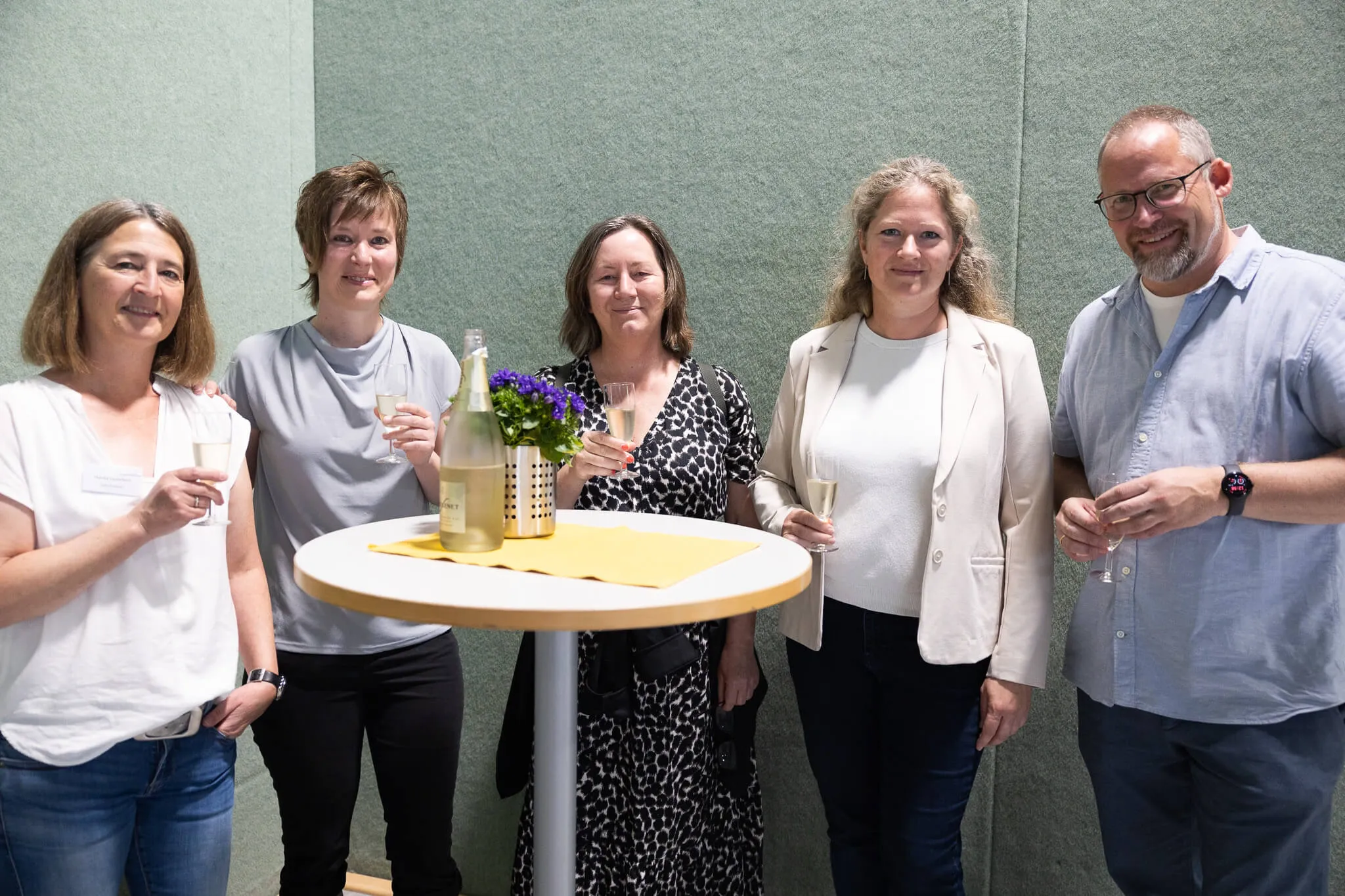 Fünf lächelnde Frauen und ein Mann stehen um einen kleinen Tisch mit einer Sektflasche und Blumen, alle halten Gläser hoch.