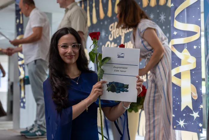 Junge Frau mit Brille und roter Rose hält eine Zeugnismappe der ESBK Halver in der Hand. Im Hintergrund sind weitere Personen und festliche Dekorationen zu sehen.