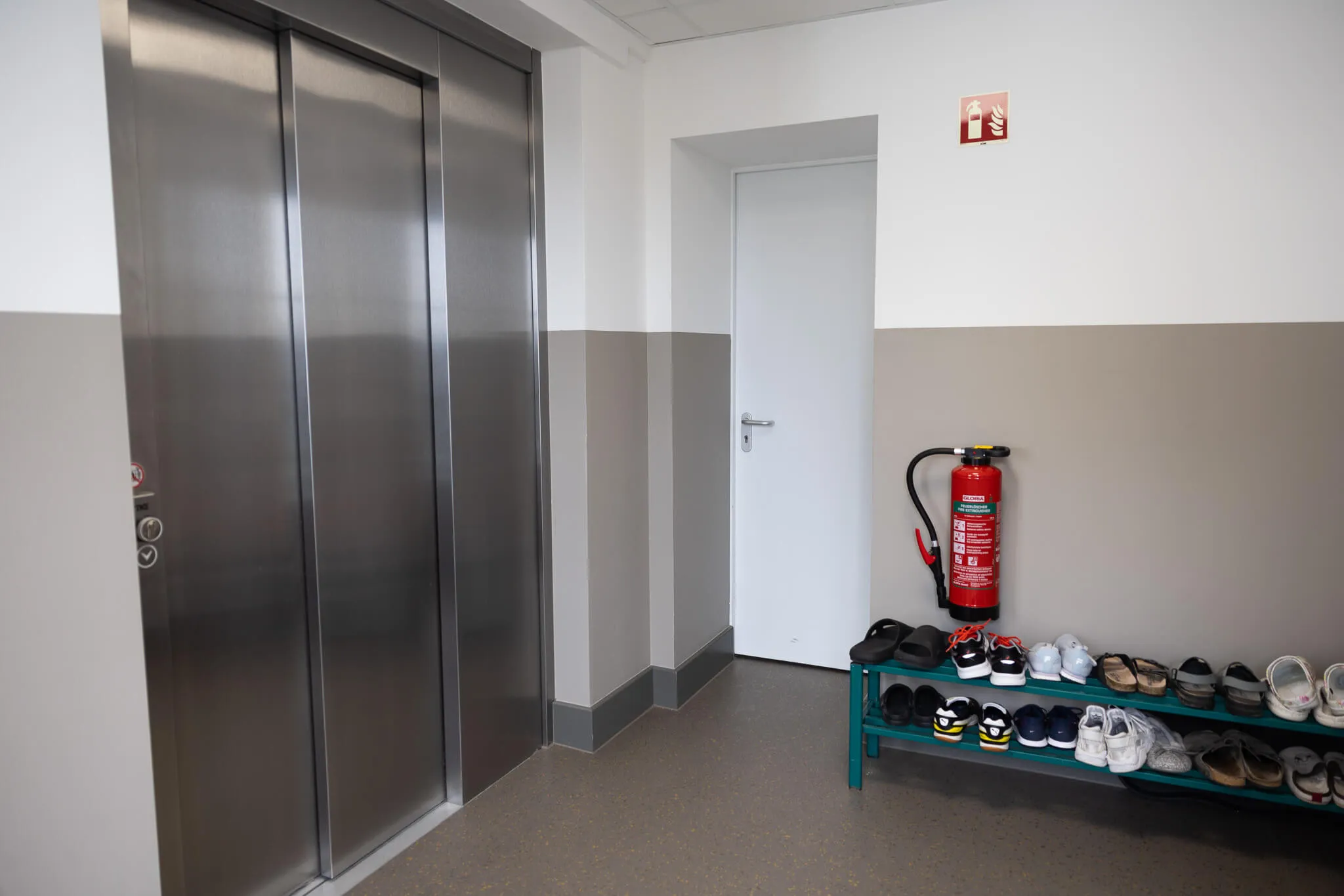 Edelstahl-Aufzugstüren neben einer Tür zu einem Raum, rechts ein Schuhregal mit mehreren Paar Schuhen und ein Feuerlöscher an der Wand.