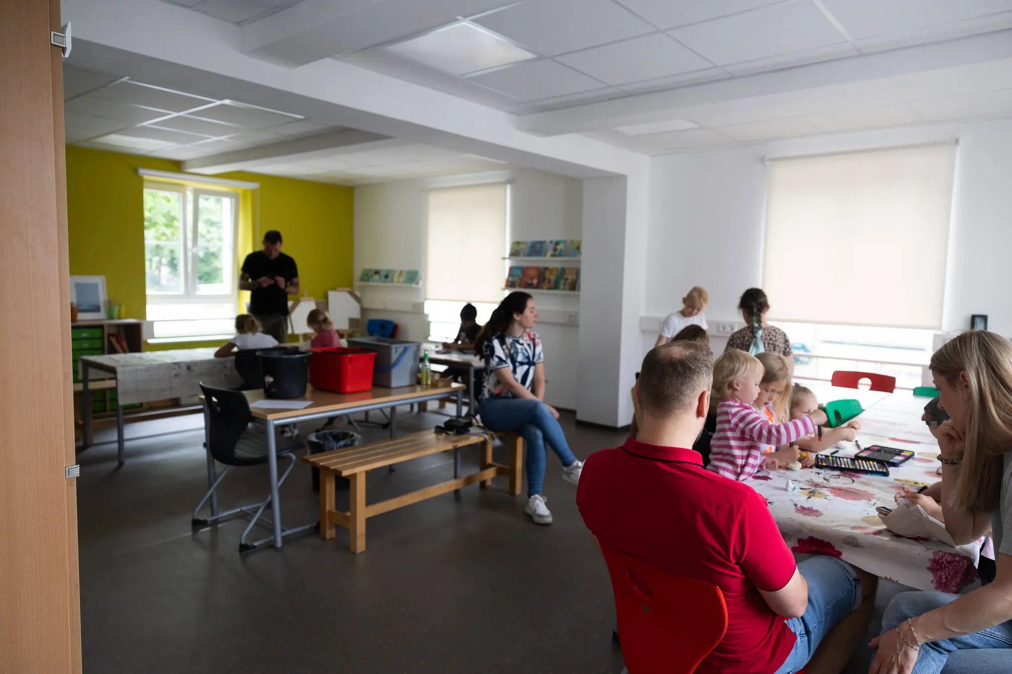 Ein heller Klassenraum mit Kindern und Erwachsenen an Tischen, während ein Mann im Hintergrund steht und spricht. Im Hintergrund ist ein Raum mit Regalen voller Bücher und Spielzeug zu sehen.