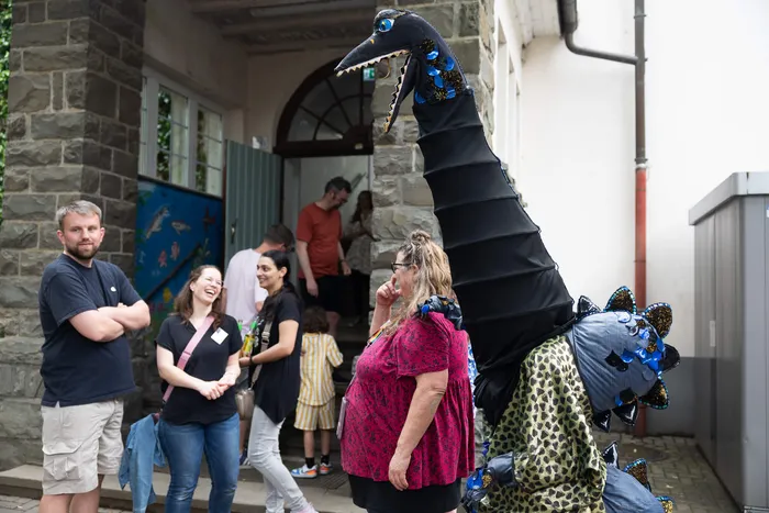 Person in einem Dinosaurierkostüm steht vor der Lindenhofschule, umgeben von feiernden Menschen.