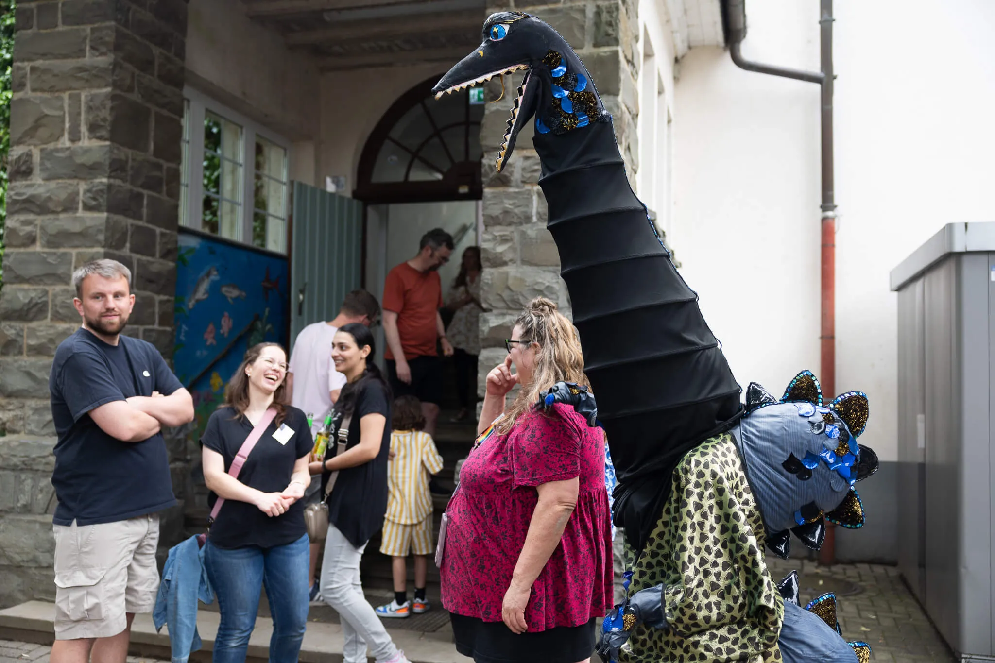 Person in einem Dinosaurierkostüm steht vor der Lindenhofschule, umgeben von feiernden Menschen.