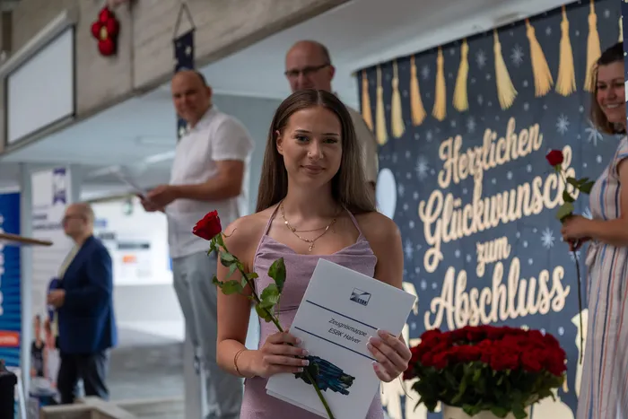 Junge Frau mit roter Rose und Zeugnismappe lächelt in die Kamera. Im Hintergrund ein Banner mit der Aufschrift „Herzlichen Glückwunsch zum Abschluss“.