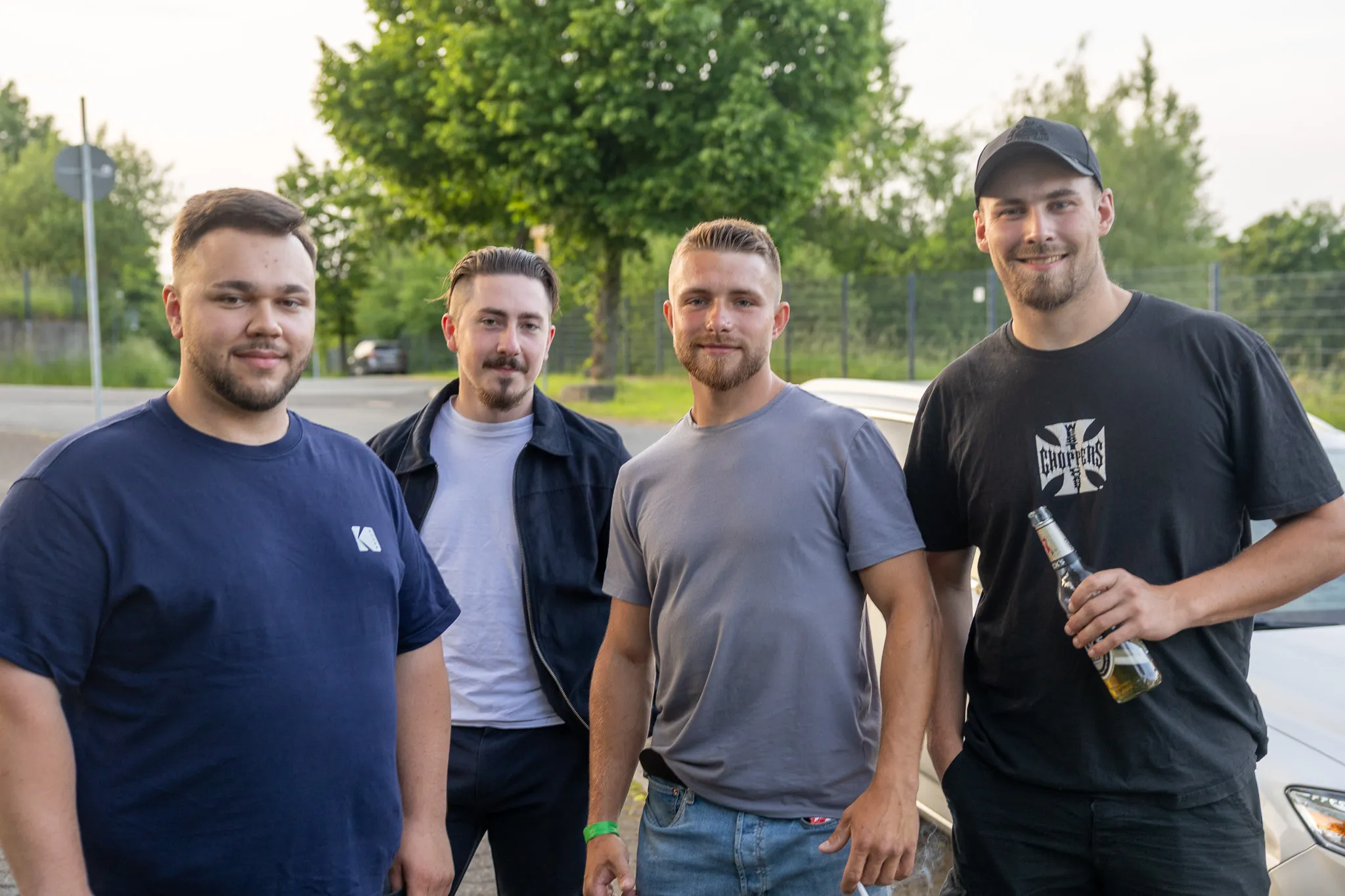 Vier junge Männer stehen lächelnd im Freien zusammen, zwei von ihnen halten Bierflaschen in der Hand.