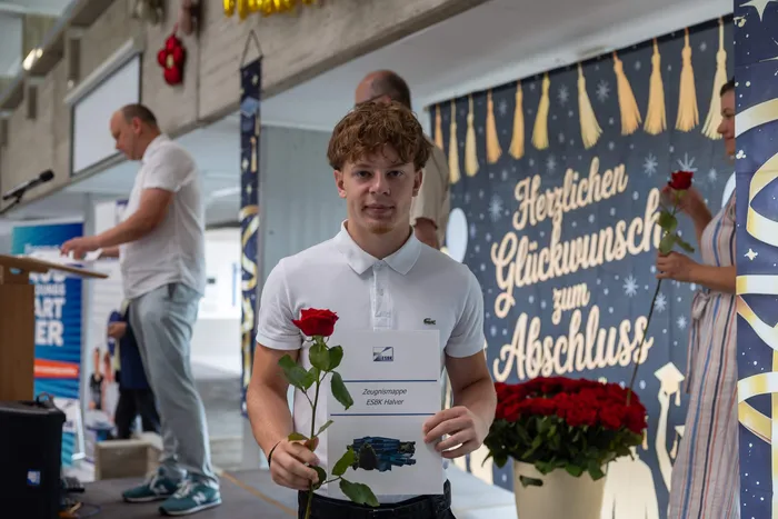 Junger Mann mit einer roten Rose und einer Zeugnismappe vor einem Hintergrund mit Glückwünschen zum Abschluss.