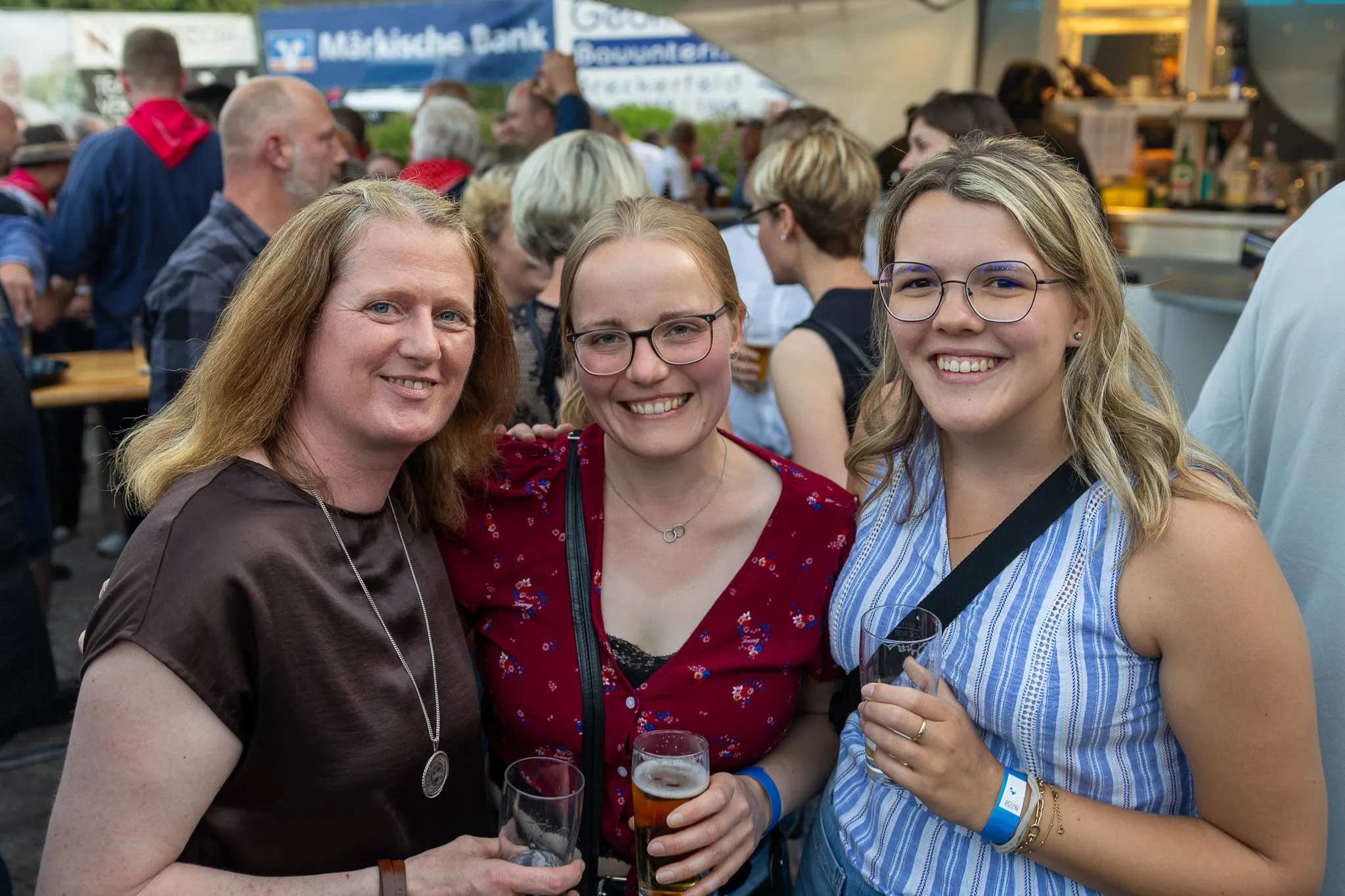 Drei lächelnde Frauen posieren im Freien mit Biergläsern in der Hand, im Hintergrund ein belebtes Festzelt.