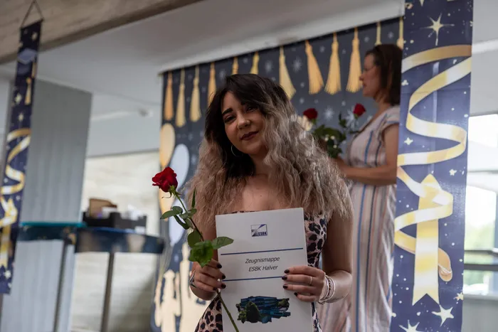 Eine junge Frau mit dunklen, lockigen Haaren hält eine rote Rose und eine Zeugnismappe der ESBK Halver. Im Hintergrund sind goldene Sterne und Vorhänge zu sehen.