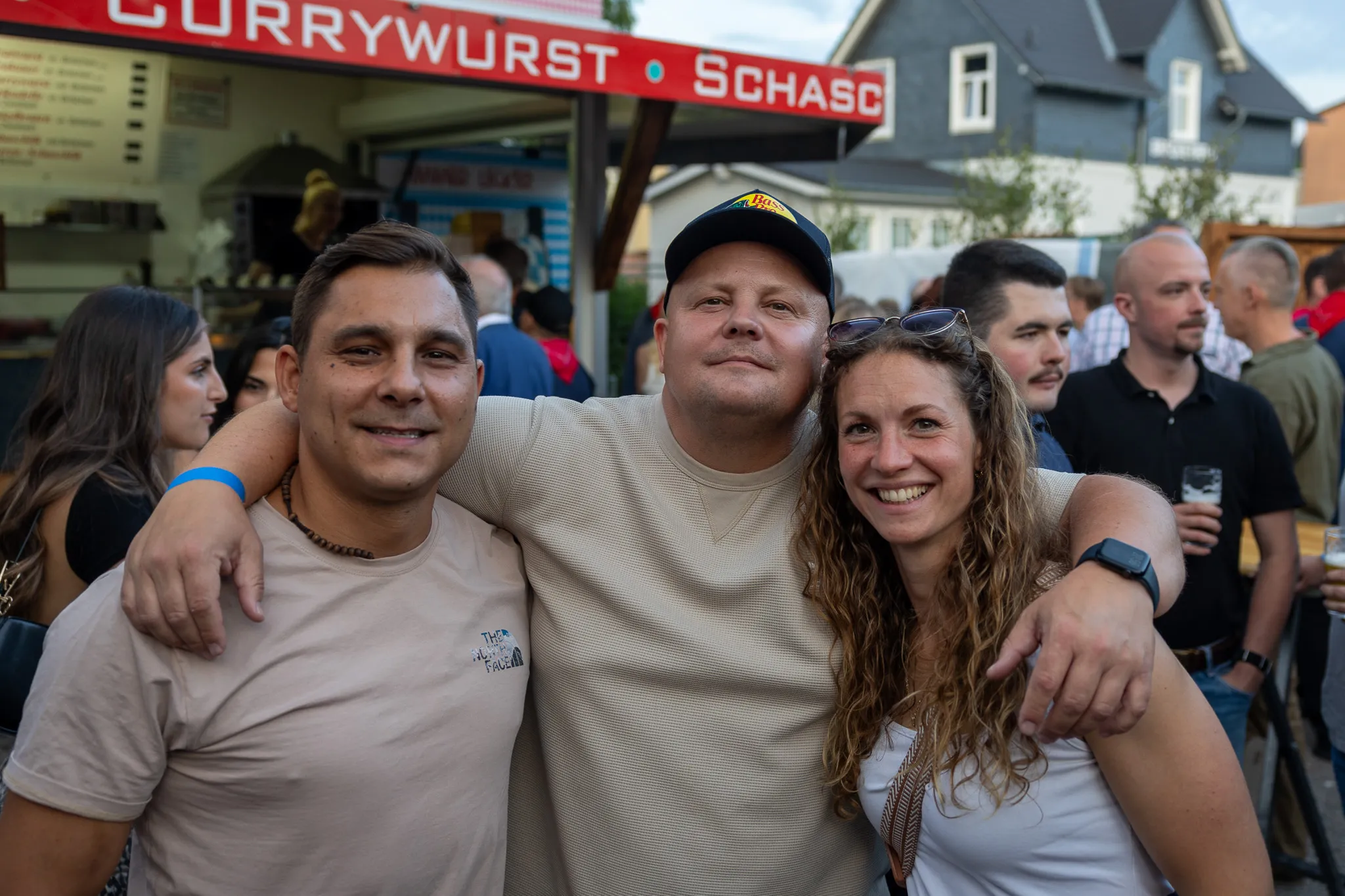 Drei lächelnde Menschen, zwei Männer und eine Frau, posieren eng umschlungen vor einer Menschenmenge und einem Imbissstand mit der Aufschrift „Currywurst“.