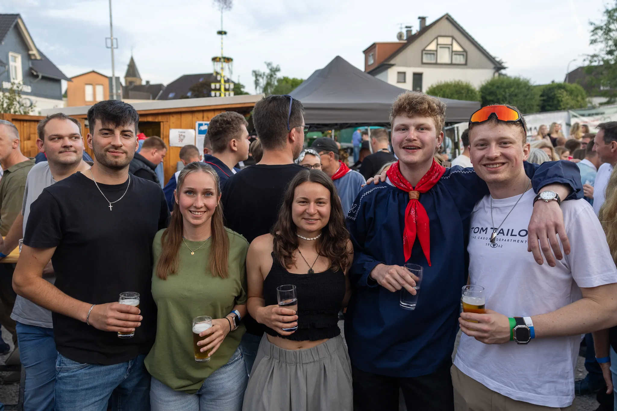 Eine Gruppe junger Erwachsener lächelt in die Kamera, während sie auf einem Volksfest Biergläser hält. Im Hintergrund sind weitere Menschen und Festzelte zu sehen.