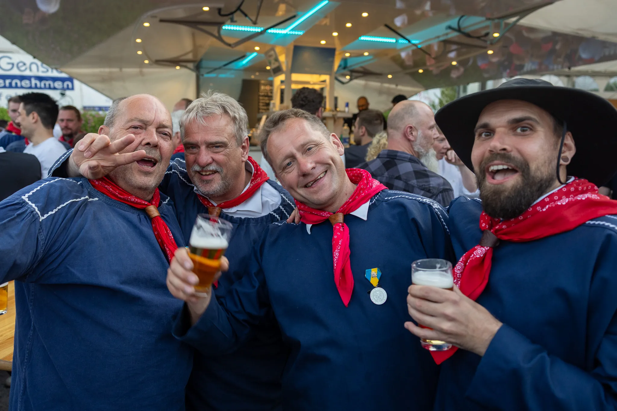 Vier Männer in traditioneller Tracht mit roten Halstüchern stoßen mit Biergläsern an und lächeln in die Kamera.