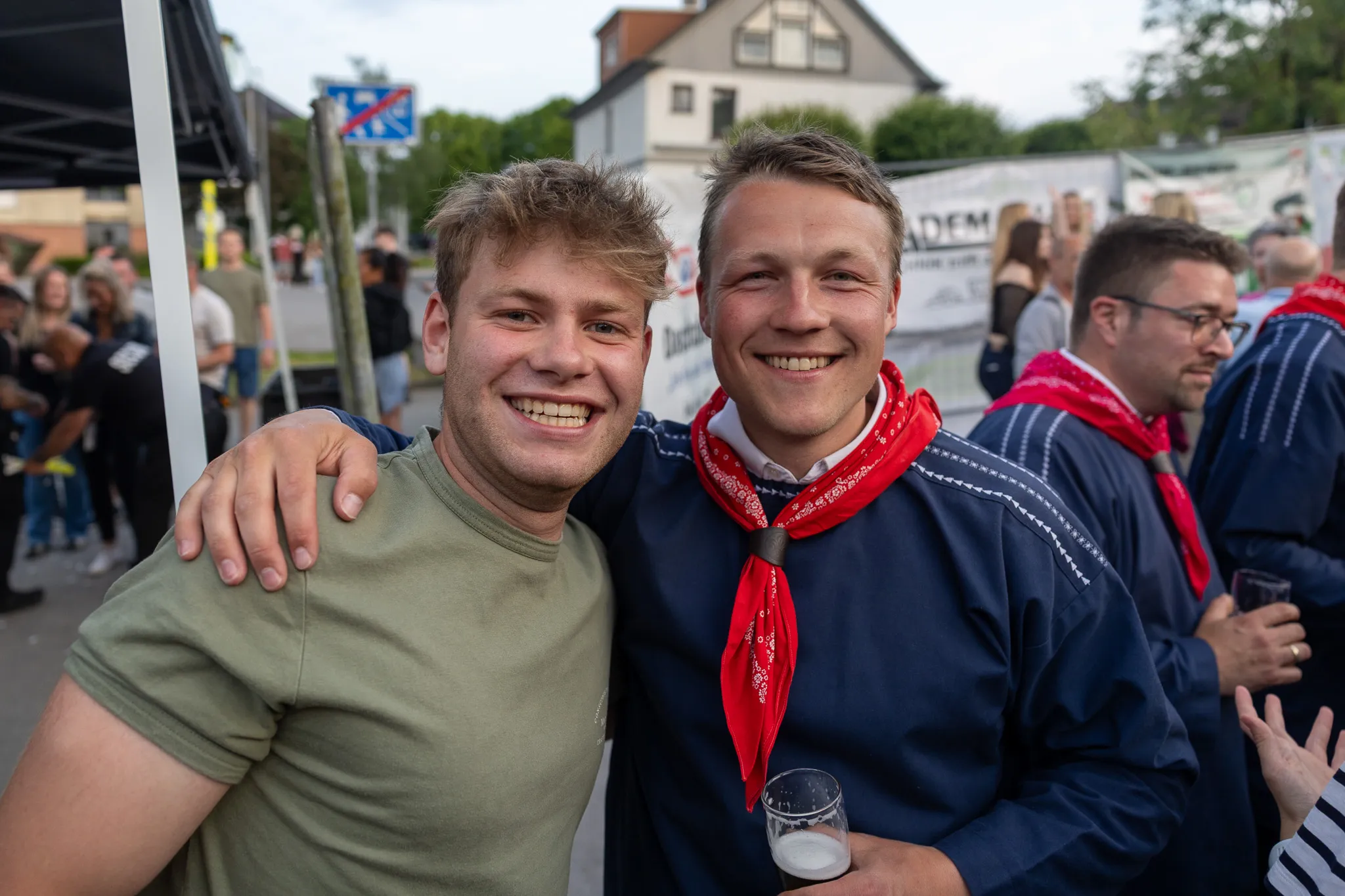 Zwei junge Männer lächeln in die Kamera, sie stehen eng zusammen und tragen traditionelle Kleidung mit roten Halstüchern. Im Hintergrund sind weitere Menschen und ein Festzelt zu sehen.