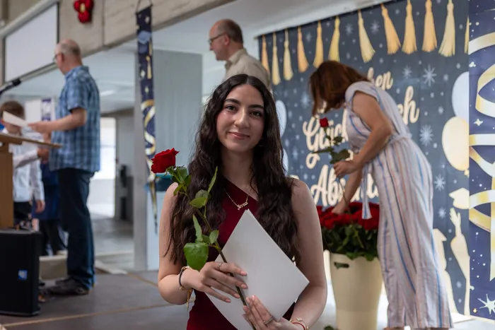 Junge Frau mit dunklen Haaren lächelt in die Kamera, hält eine rote Rose und eine Urkunde. Im Hintergrund eine Frau und ein Mann sowie ein Banner mit der Aufschrift „Glück Auf“.