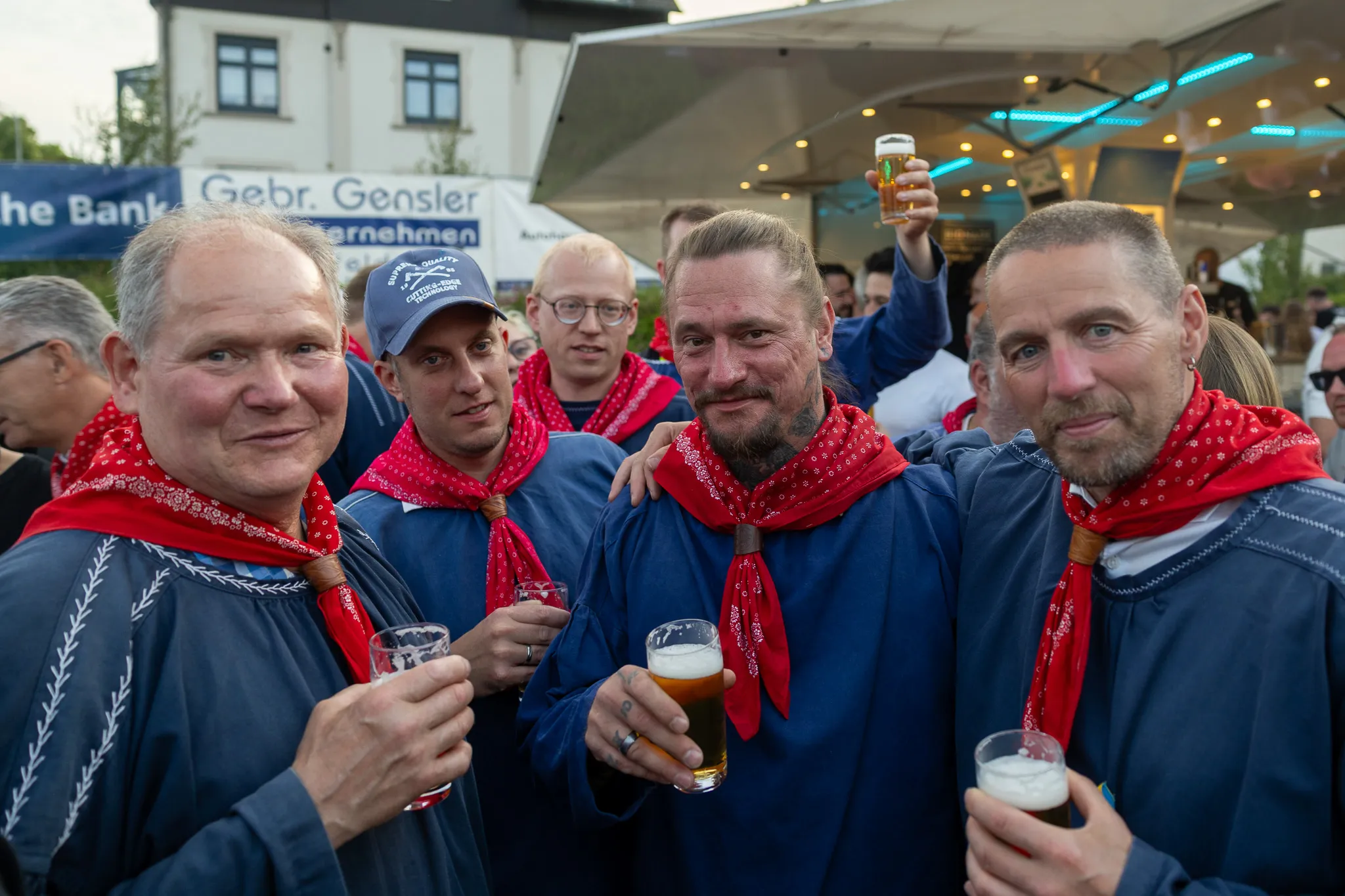 Vier Männer mit roten Halstüchern und traditioneller Tracht stoßen mit Biergläsern an, im Hintergrund ein Festzelt und weitere feiernde Menschen.