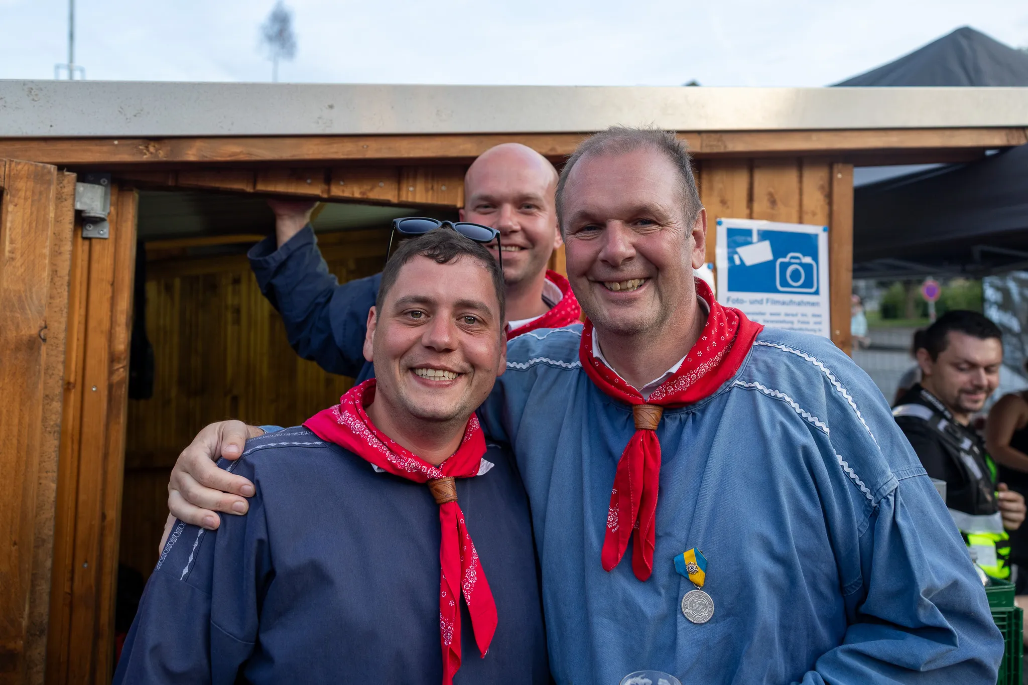 Zwei Männer in traditioneller Tracht mit roten Halstüchern lächeln in die Kamera, ein dritter Mann steht im Hintergrund. Im Hintergrund ist ein Schild mit einem Kamera-Symbol zu sehen.