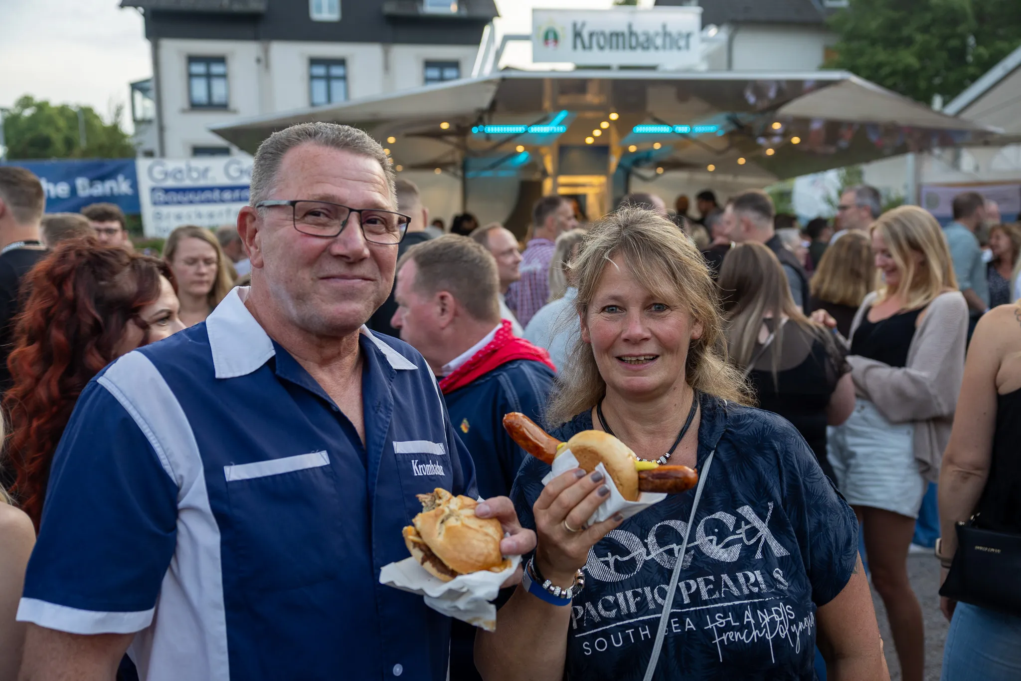 Zwei lächelnde Menschen, ein Mann mit Brille und eine Frau, halten jeweils ein belegtes Brötchen und eine Bratwurst in der Hand, umgeben von Menschen in einem belebten Festzelt.