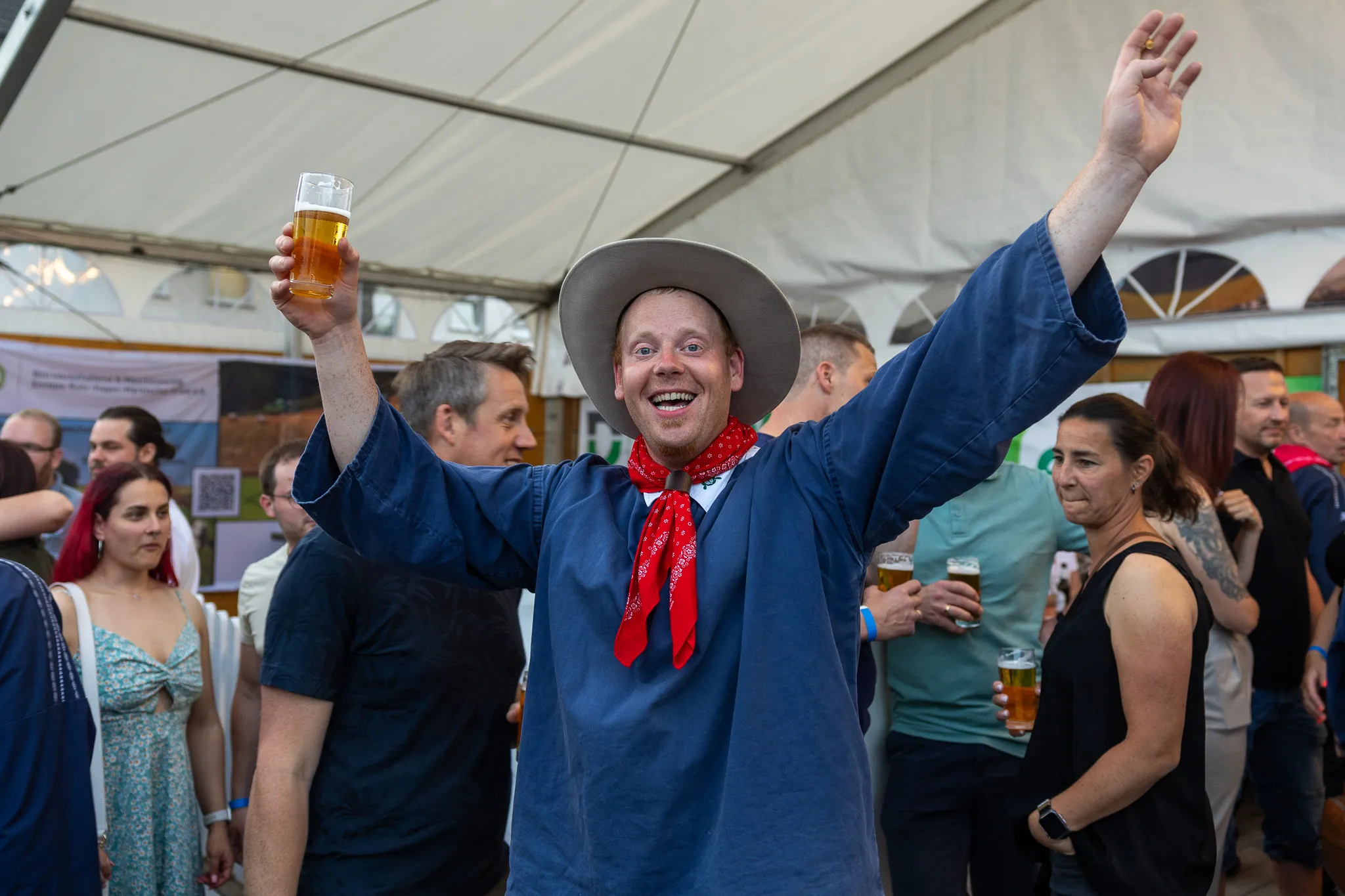 Ein Mann in Tracht mit Cowboyhut hält ein Bier hoch und winkt in einem vollen Festzelt.