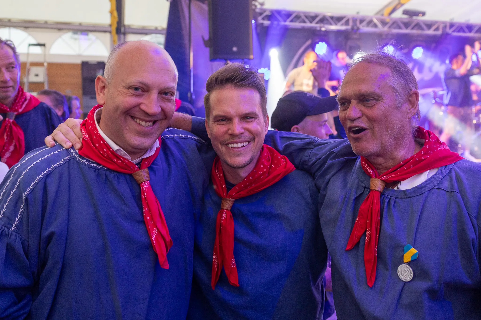 Drei lächelnde Männer in traditioneller blau-weißer Tracht mit roten Halstüchern posieren eng zusammen vor einer Bühne mit Musikern.