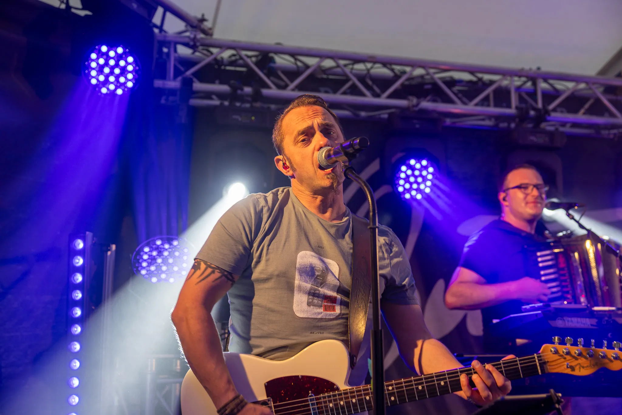 Sänger mit Gitarre performt auf einer Bühne vor Publikum, während ein Musiker im Hintergrund ein Akkordeon spielt.