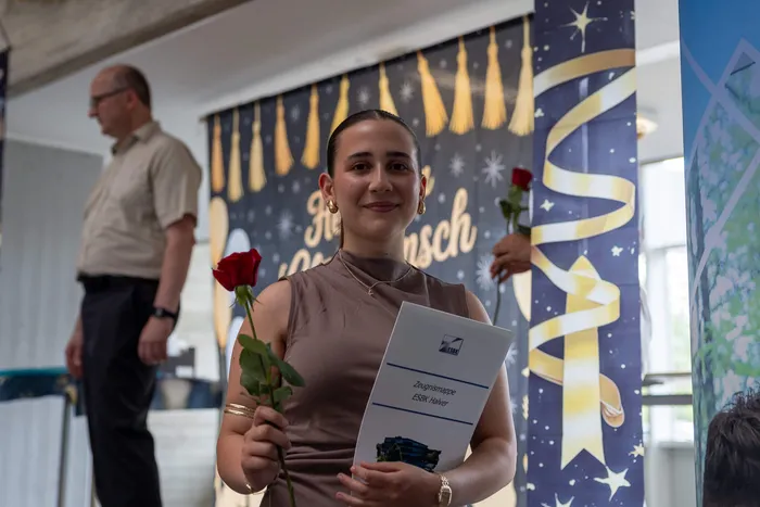 Eine junge Frau mit dunklen Haaren nimmt im Rahmen einer Abschlussfeier eine Urkunde und zwei rote Rosen entgegen. Im Hintergrund steht ein Mann und ein Banner mit dem Schriftzug „Absolventen“.