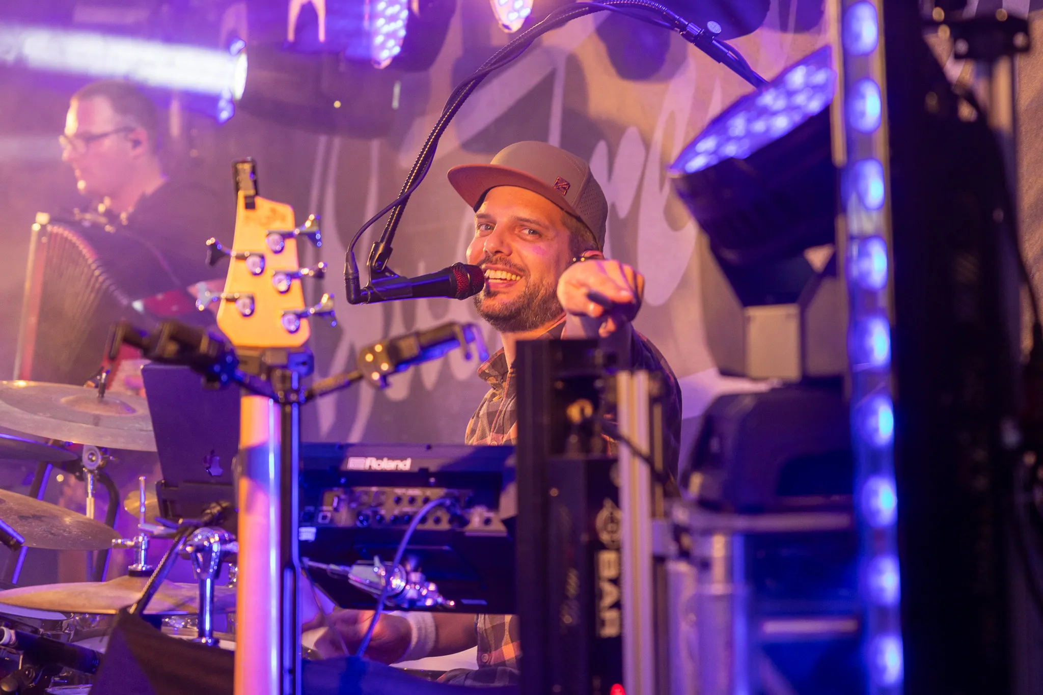 Sänger mit Cap und Mikrofon performt vor Bandkollegen auf der Bühne, im Hintergrund Lichteffekte und Instrumente.