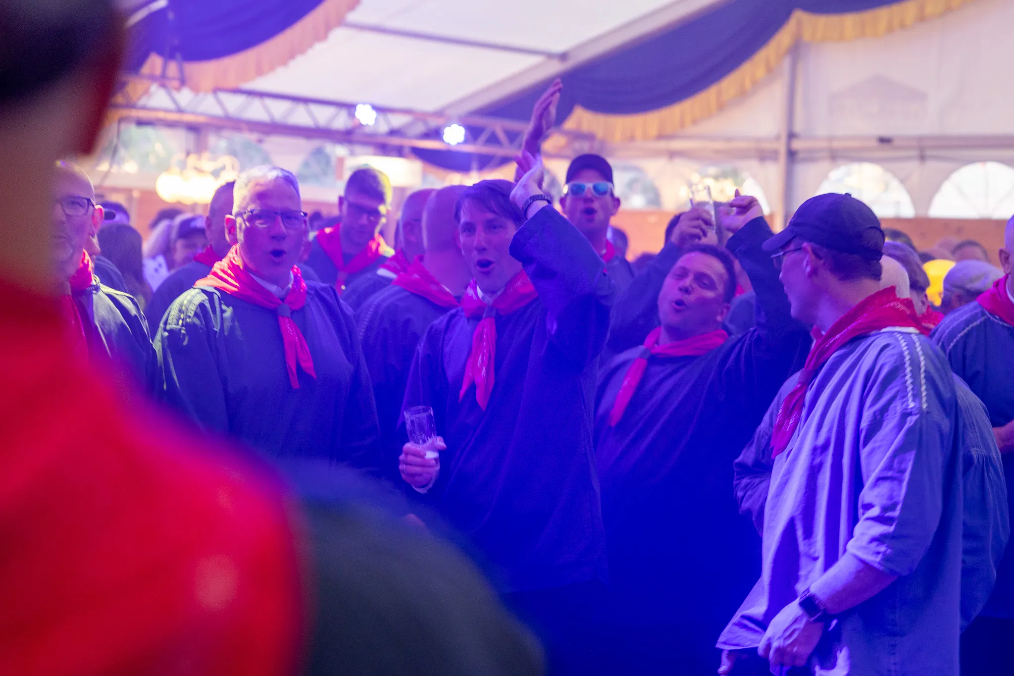 Mehrere Männer und Frauen singen und feiern im Festzelt, viele tragen rote Halstücher. Im Hintergrund ist ein Zelt mit Lichterketten zu sehen.