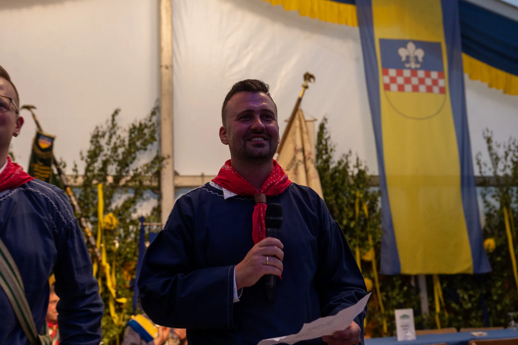 Ein Mann hält ein Mikrofon und spricht in einem Festzelt, im Hintergrund eine Flagge mit blauem und gelbem Wappen.