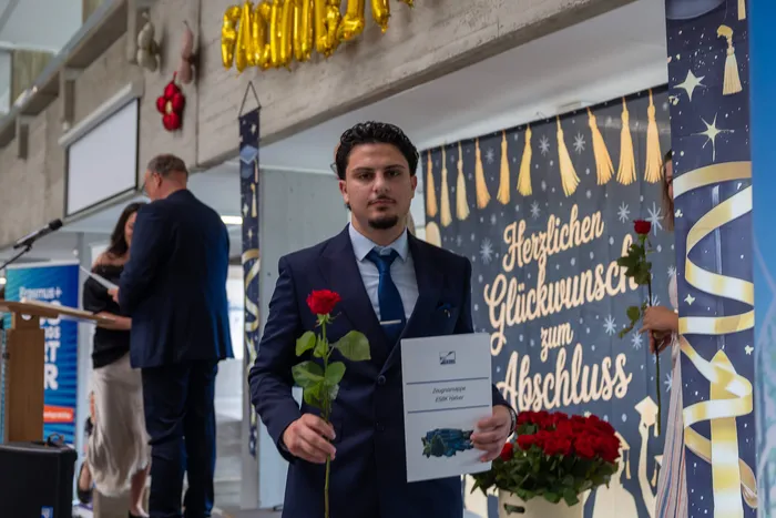 Ein junger Mann in Anzug und Krawatte hält eine rote Rose und eine Urkunde in der Hand, vor ihm ein Banner mit der Aufschrift „Herzlichen Glückwunsch zum Abschluss“.