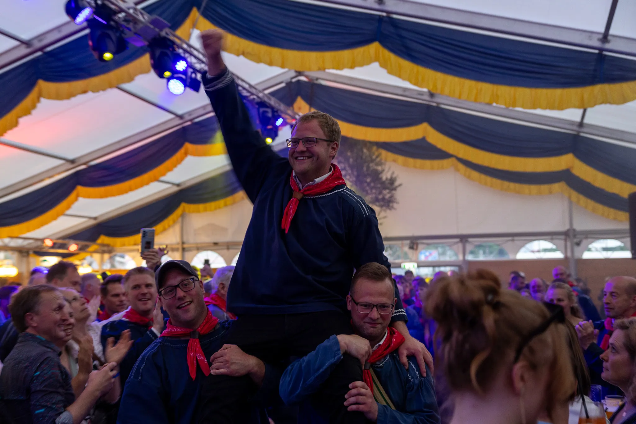 Ein Mann wird auf den Schultern von zwei anderen Männern durch ein Festzelt getragen, während umstehende Menschen applaudieren und jubeln. Im Hintergrund sind gelbe und blaue Stoffbahnen an der Zeltdecke zu sehen.