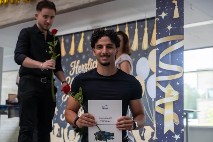 Ein junger Mann mit dunklen lockigen Haaren lächelt in die Kamera und hält eine Urkunde sowie eine rote Rose. Im Hintergrund steht ein weiterer junger Mann und ein Banner mit goldenen Quasten.