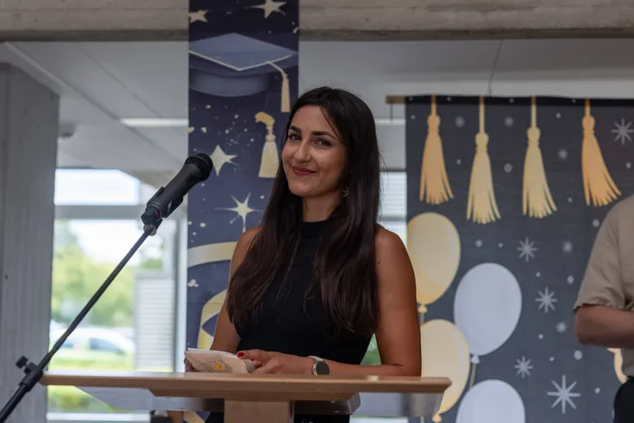 Eine Frau mit langen, dunklen Haaren lächelt in die Kamera, während sie an einem Rednerpult steht und eine Rede hält. Im Hintergrund ist ein Banner mit Hüten und Sternen zu sehen.