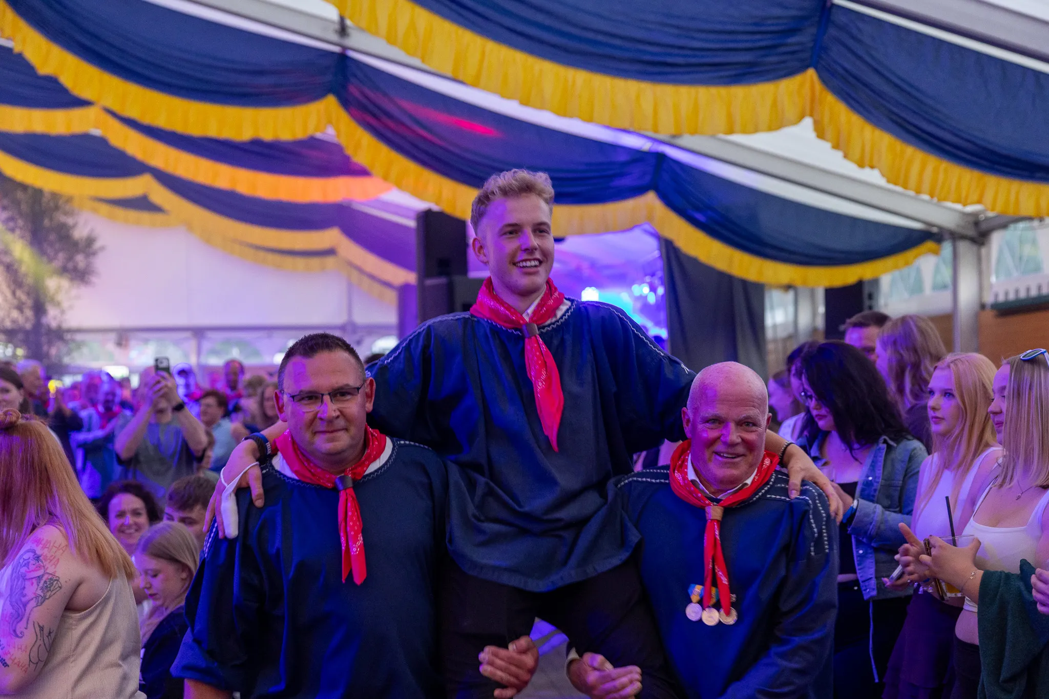 Drei Männer in traditioneller Tracht werden auf den Schultern getragen, umgeben von feiernden Menschen in einem Festzelt.
