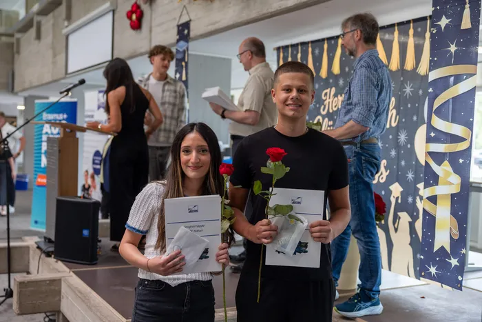 Zwei junge Absolventen, eine Frau und ein Mann, halten Urkunden und jeweils eine rote Rose in den Händen. Im Hintergrund stehen weitere Personen und eine festliche Dekoration.