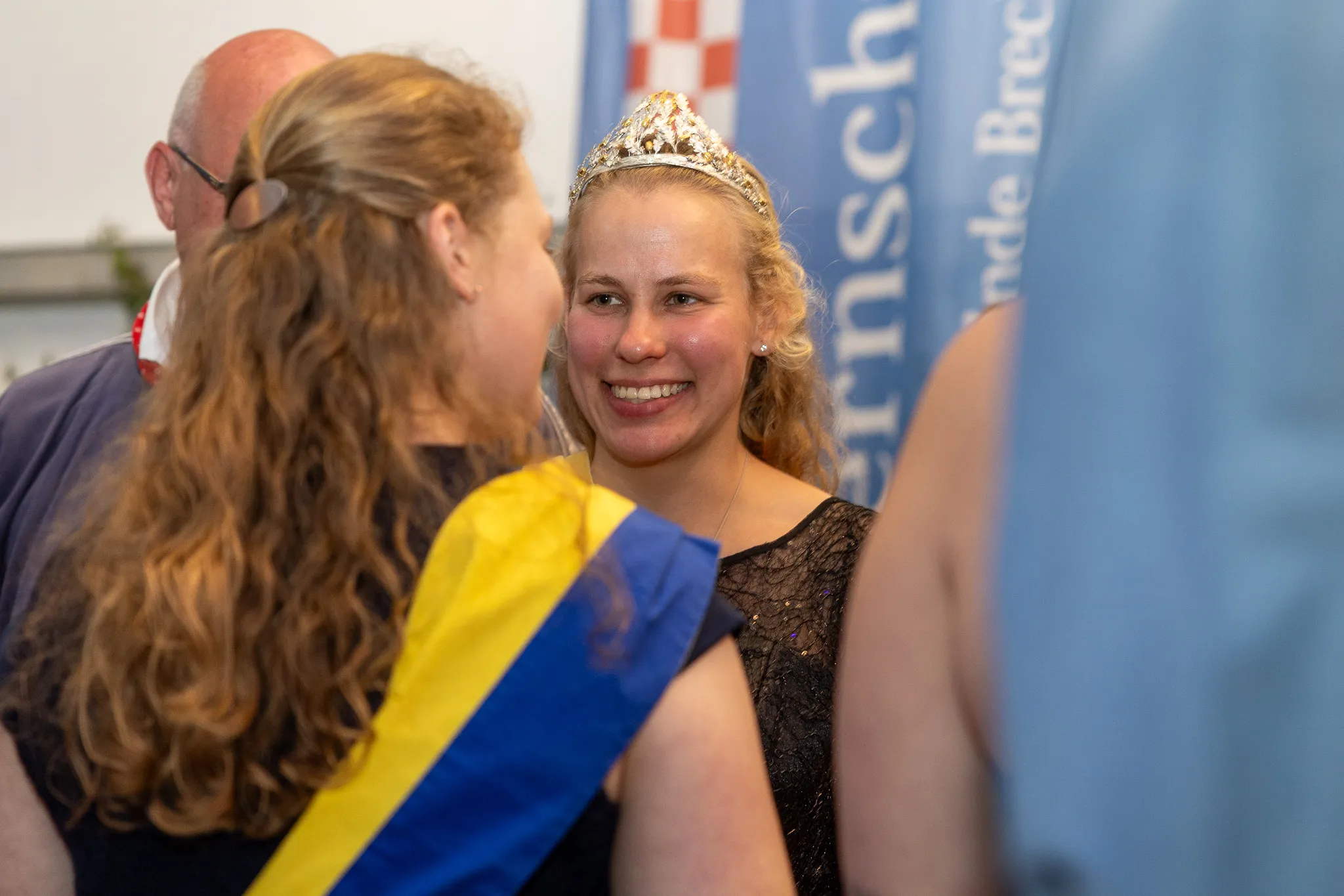 Junge Frau mit Tiara und blauem Band lächelt in die Kamera, während eine andere Person ihr etwas ins Ohr flüstert. Im Hintergrund ein Banner mit dem Schriftzug „Bauernschützen“.
